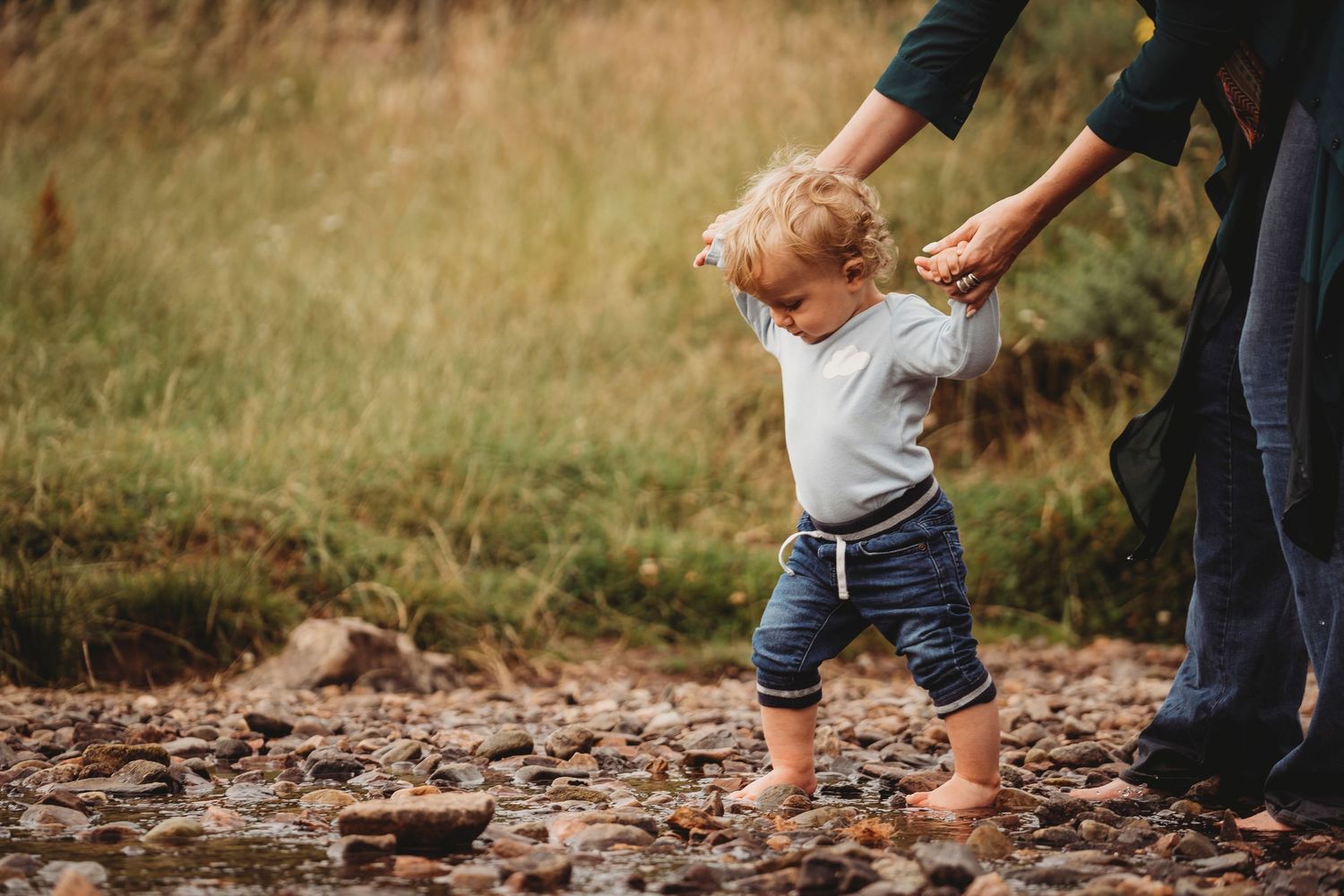 A toddler takes first steps on rocky ground while an adult holds their hands for support outdoors.