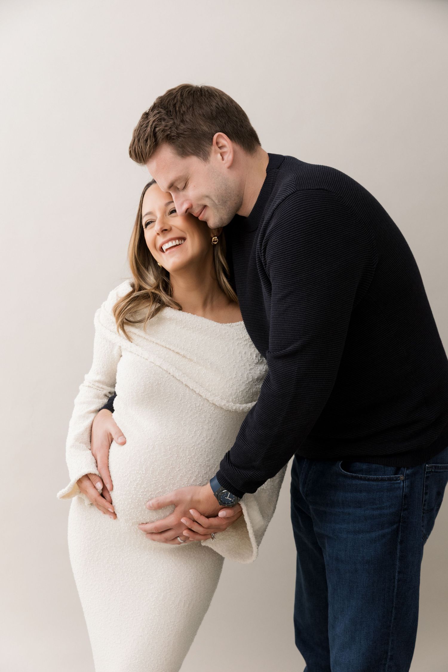 A serene maternity moment by a bright window in winter, wearing a cozy white sweater dress against natural light.