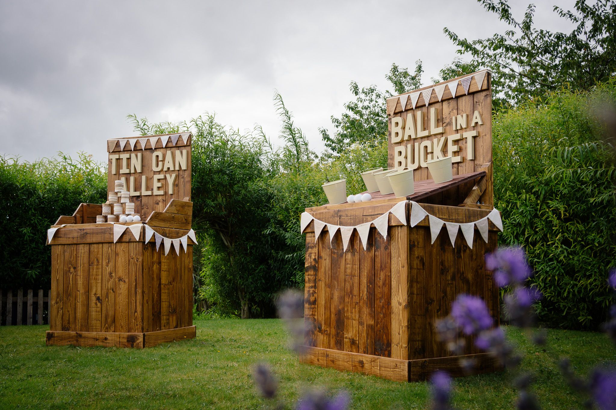 A rustic wooden carnival game booth with bunting flags sits in a lush green garden setting.