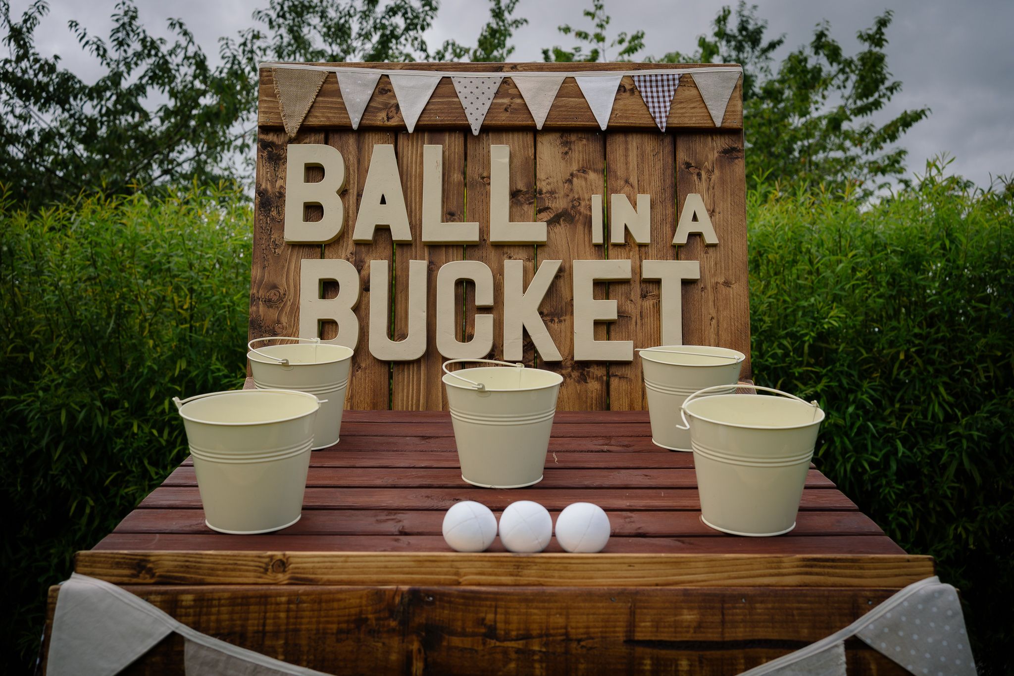 Rustic wooden carnival game setup with 'Ball in a Bucket' sign, white buckets and balls on display table.