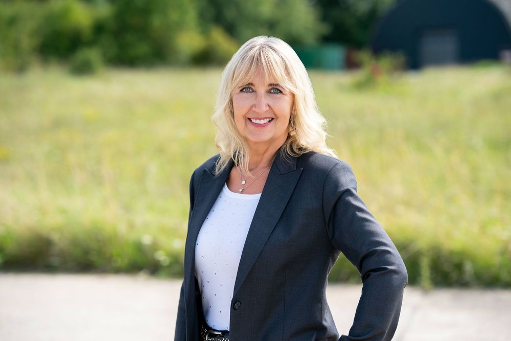 Professional wearing a black blazer and white top stands outdoors in a sunny field.