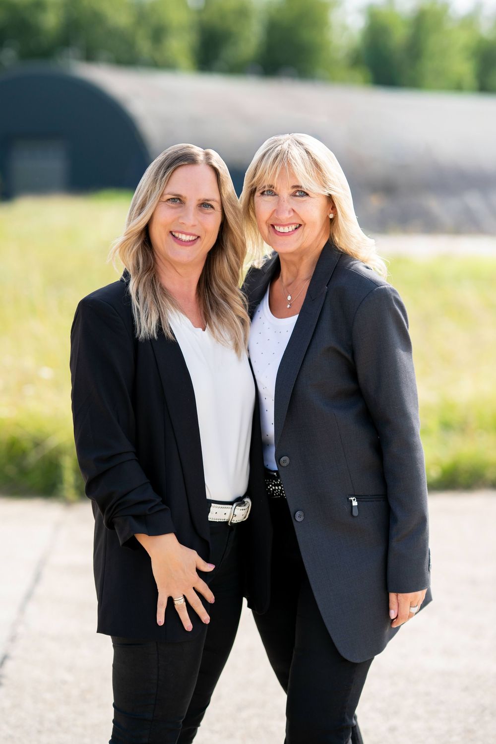 Two professional women in business attire stand together outdoors smiling on a sunny day.