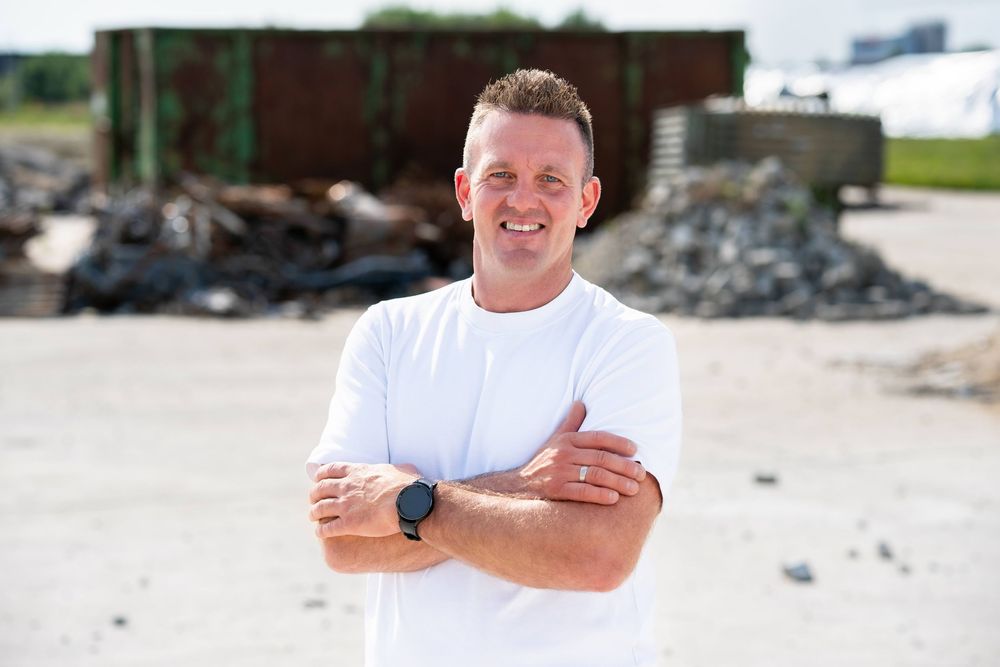 A person in a white t-shirt stands with crossed arms and a blue watch in front of an industrial recycling yard.
