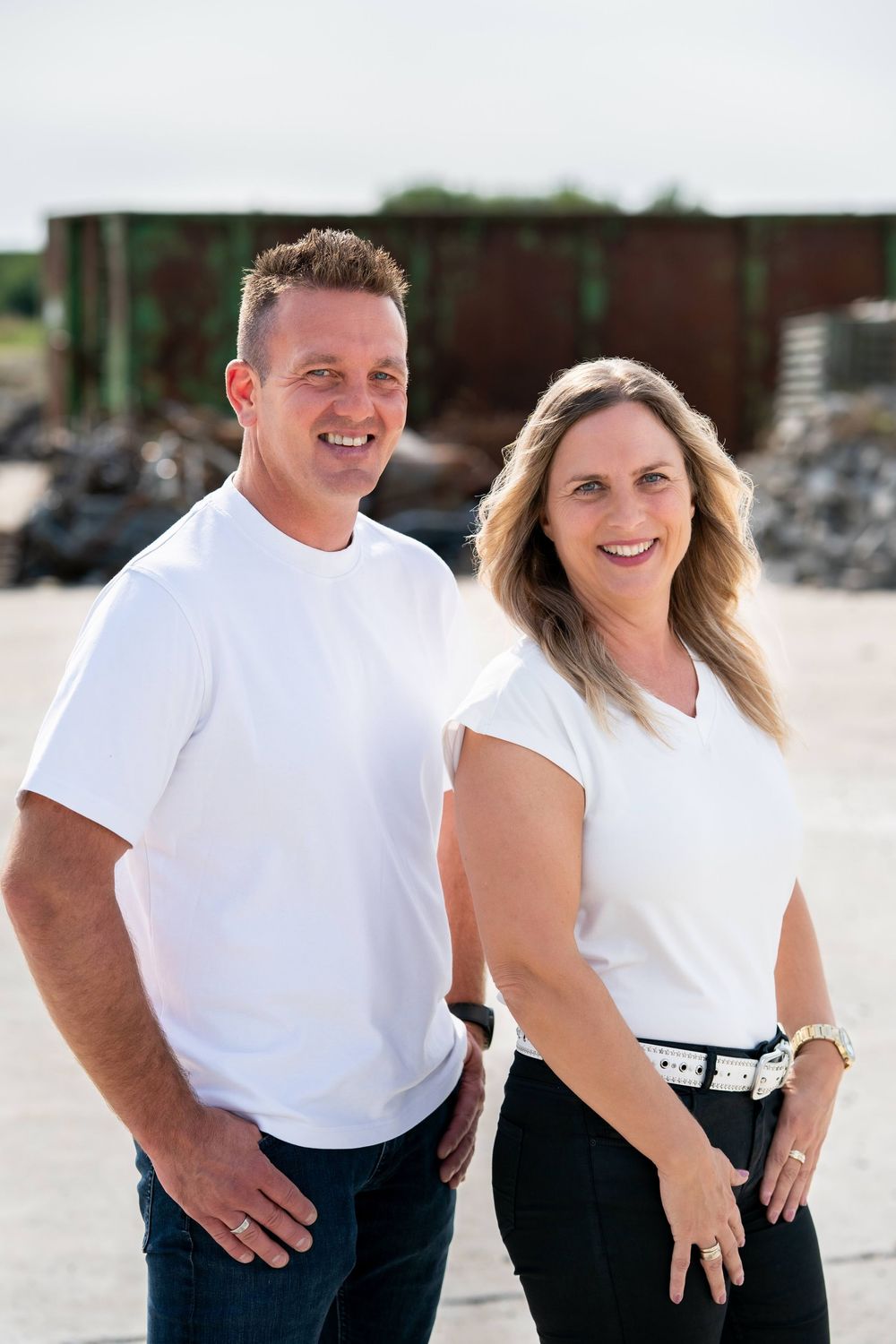 A couple in matching white t-shirts and dark jeans smiling together outdoors in an industrial setting.