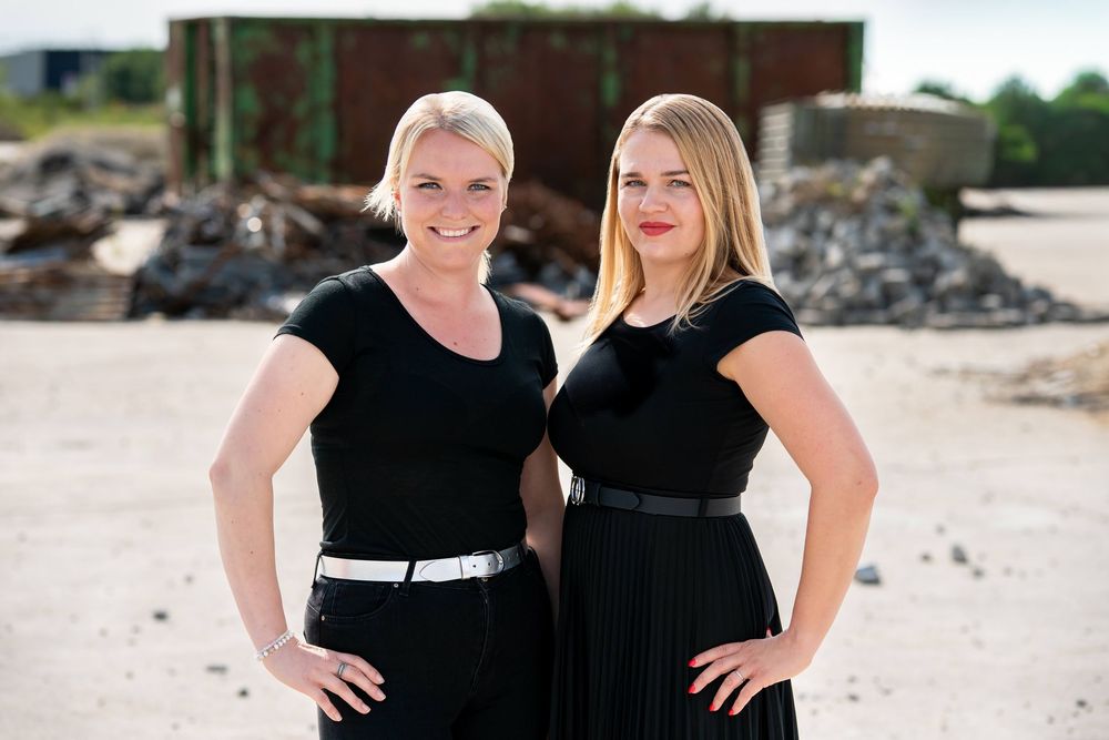 Two women in black dresses pose together for photos at an outdoor beach location.