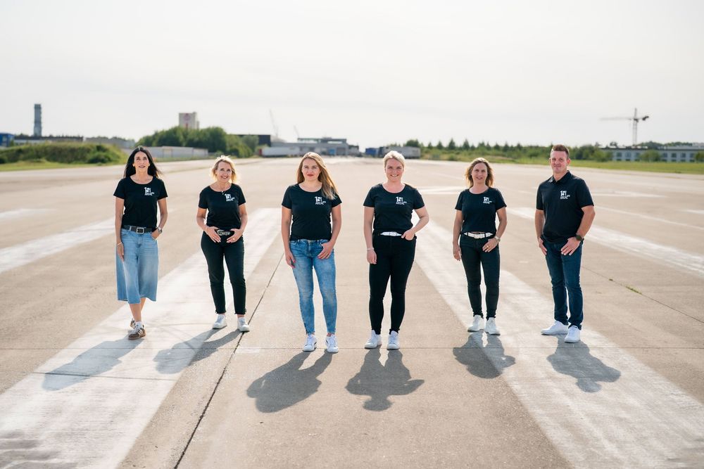 Six people wearing matching black shirts stand in a row on concrete ground outdoors.