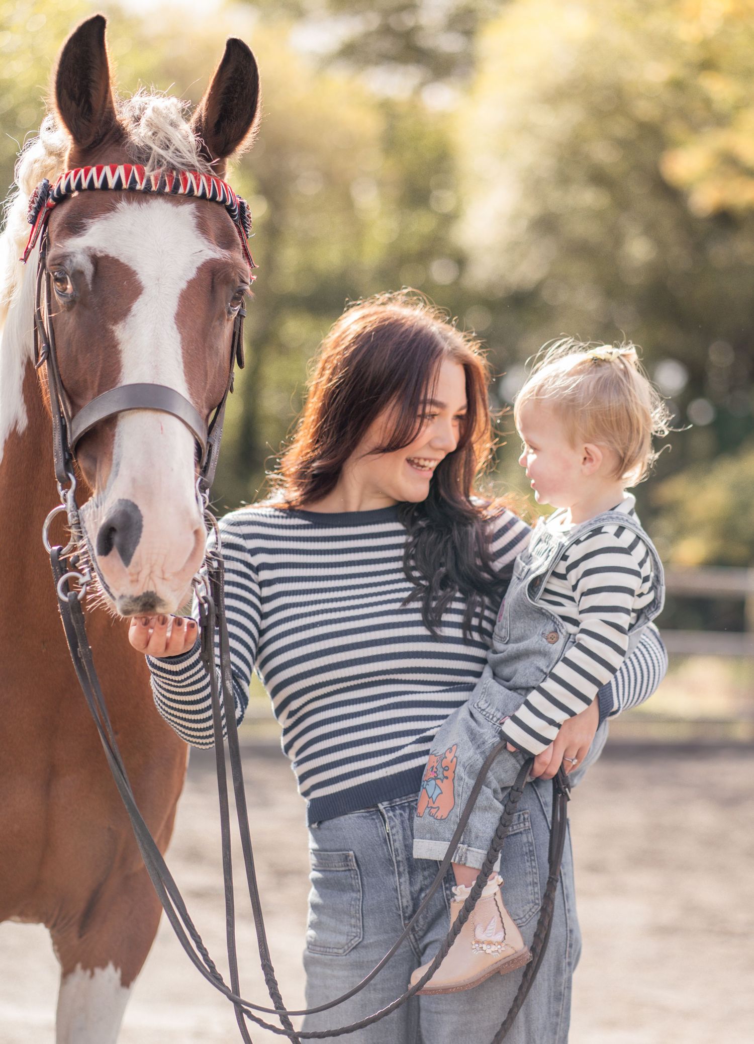 Parent and child in matching striped shirts share tender moments with a brown and white horse in outdoor setting.
