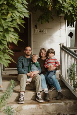 A family sits together on their front porch steps with green foliage framing the scene.