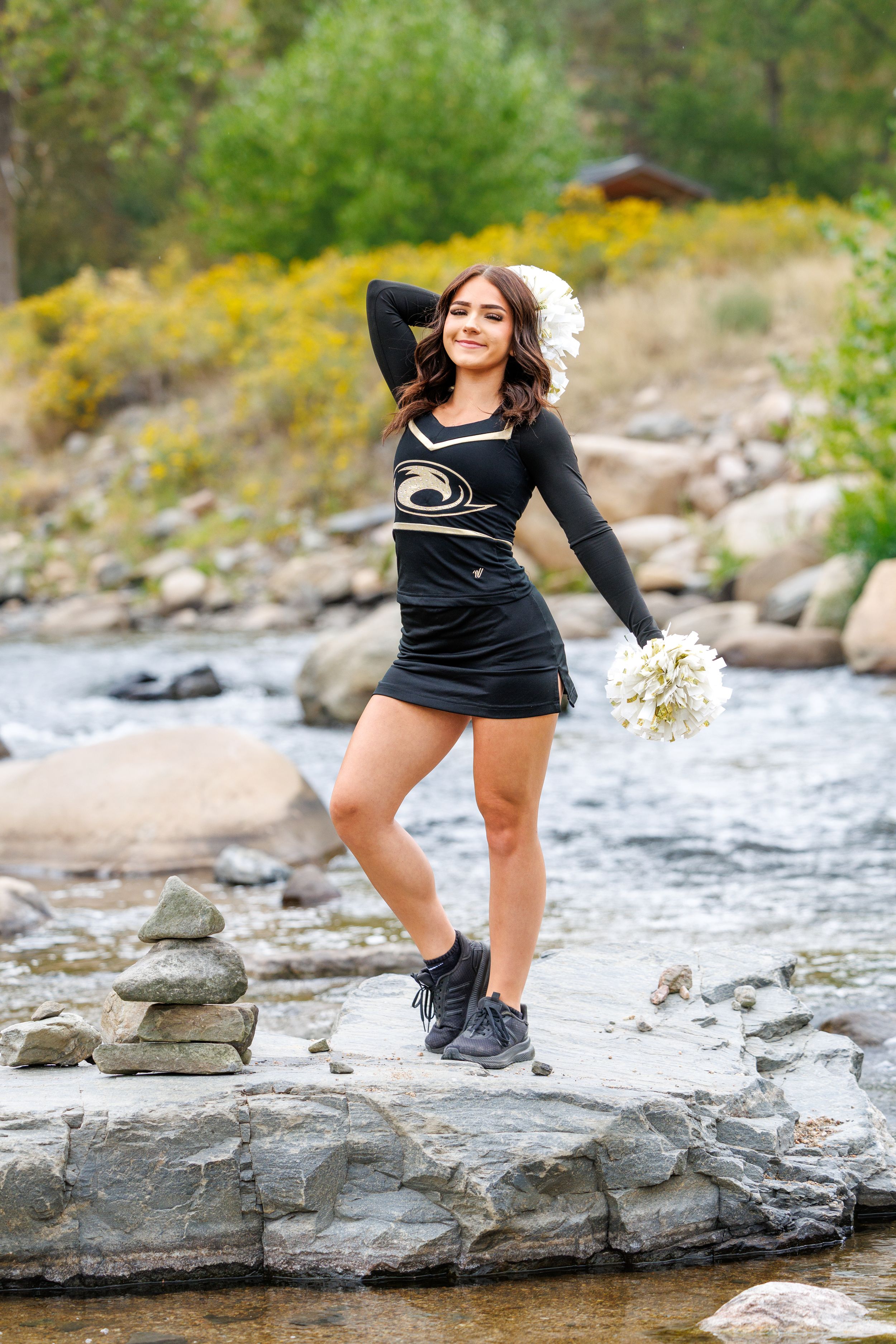 Cheerleader in black uniform holds pom-pom while posing on rocks by a mountain stream.