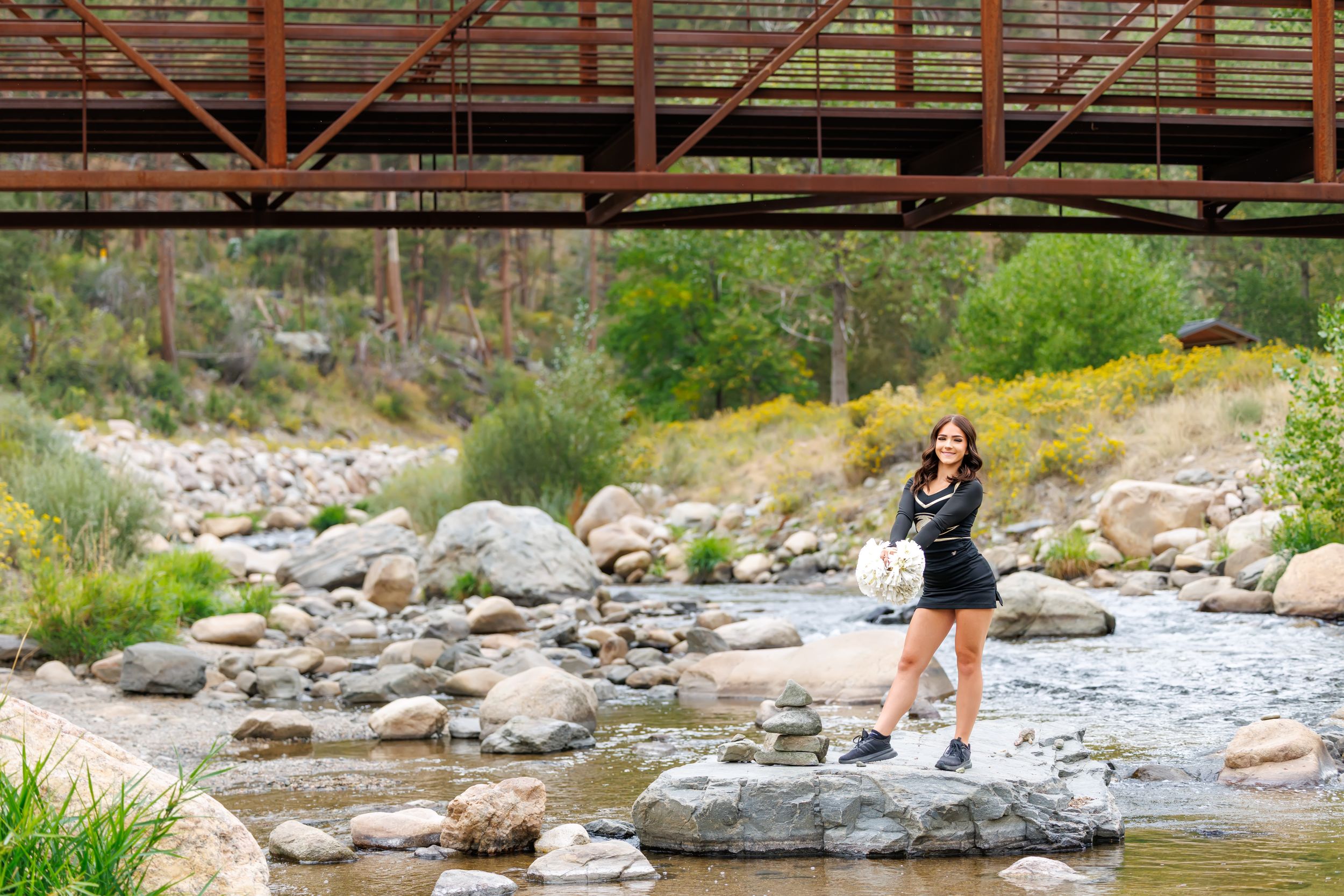 A young woman stands on rocks by a shallow creek beneath a rustic bridge surrounded by lush greenery.