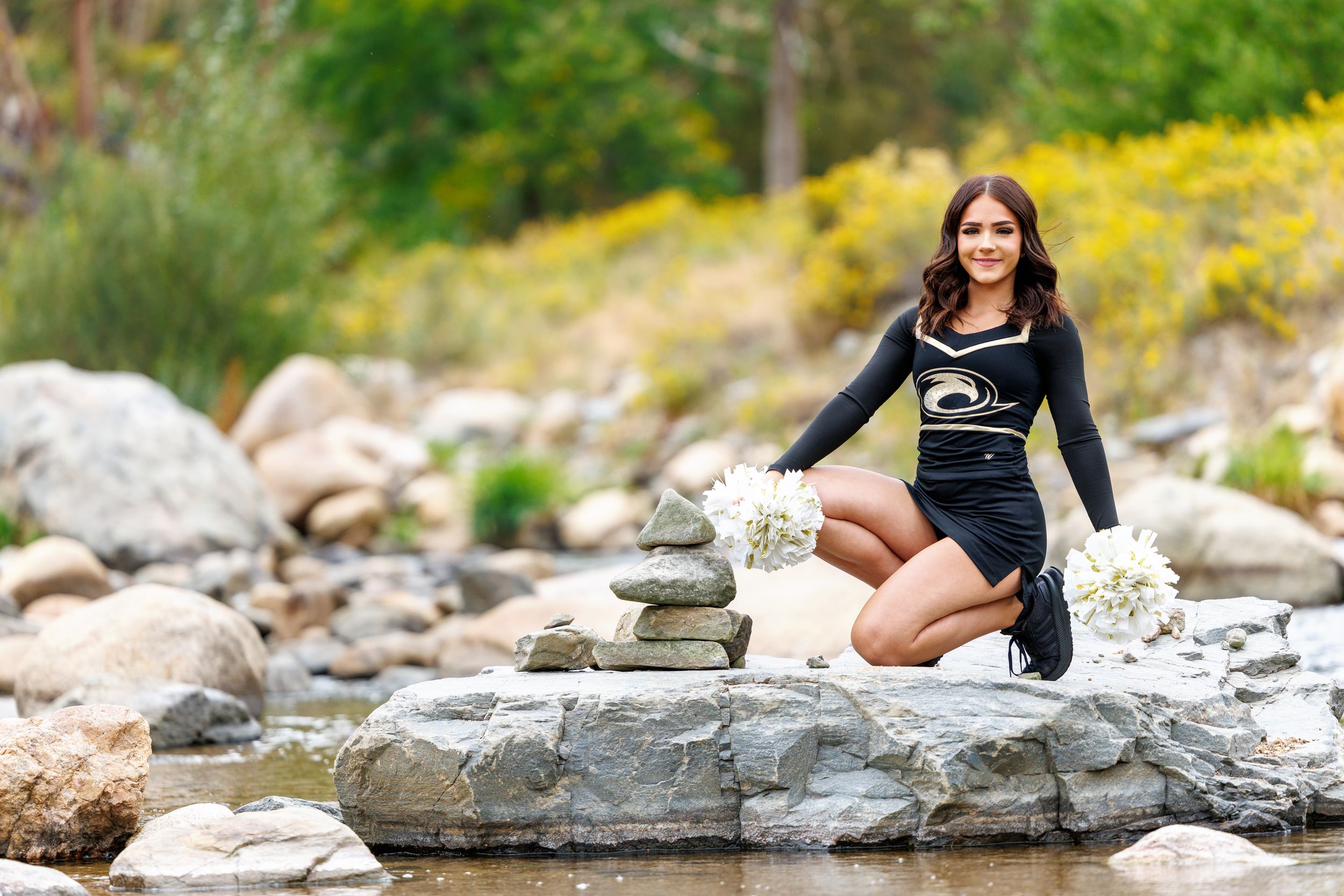 Cheerleader in black uniform poses on rocks by a stream with white pom-poms in a scenic outdoor setting.