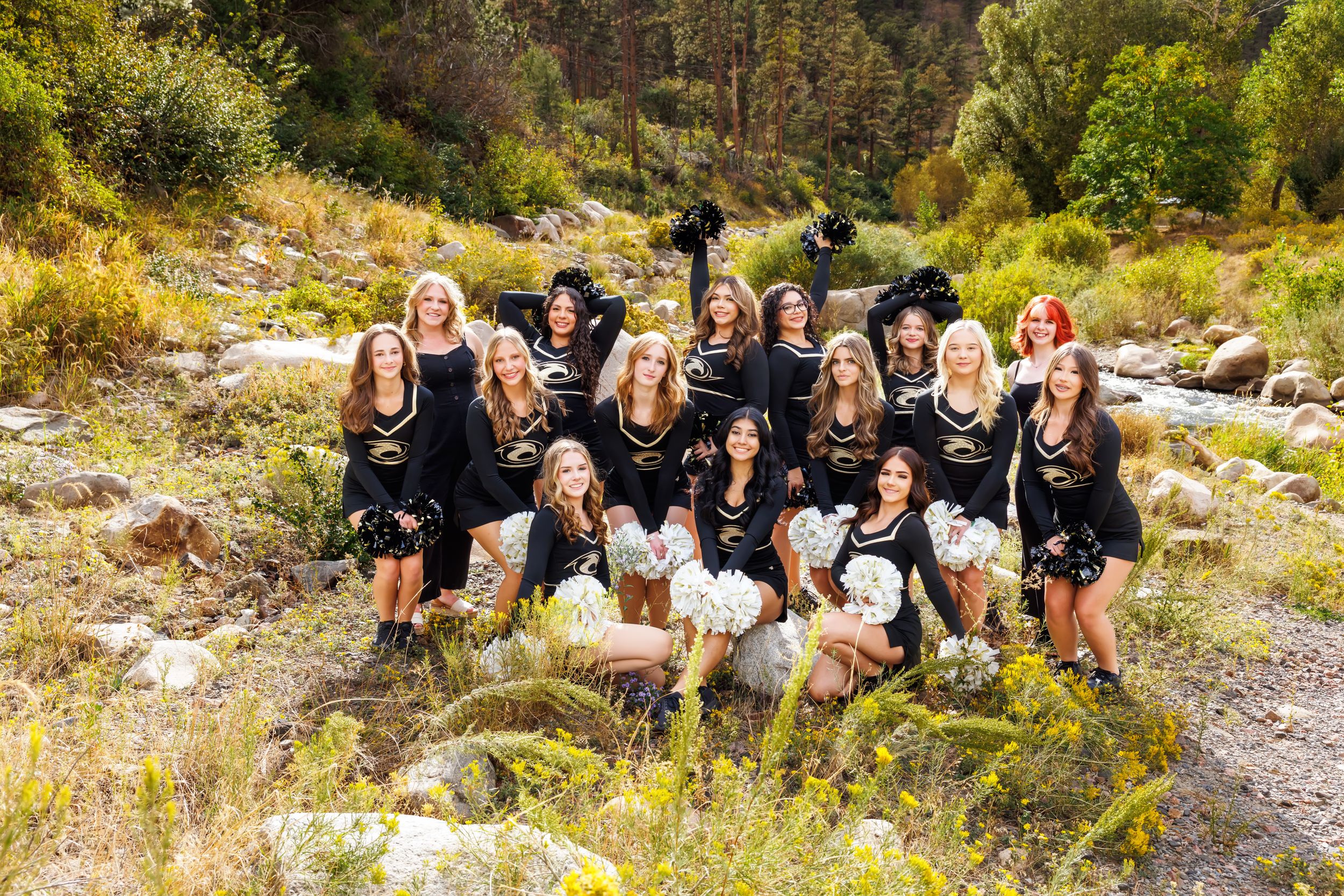 Group of cheerleaders in black uniforms posing outdoors near a rocky stream surrounded by lush greenery.