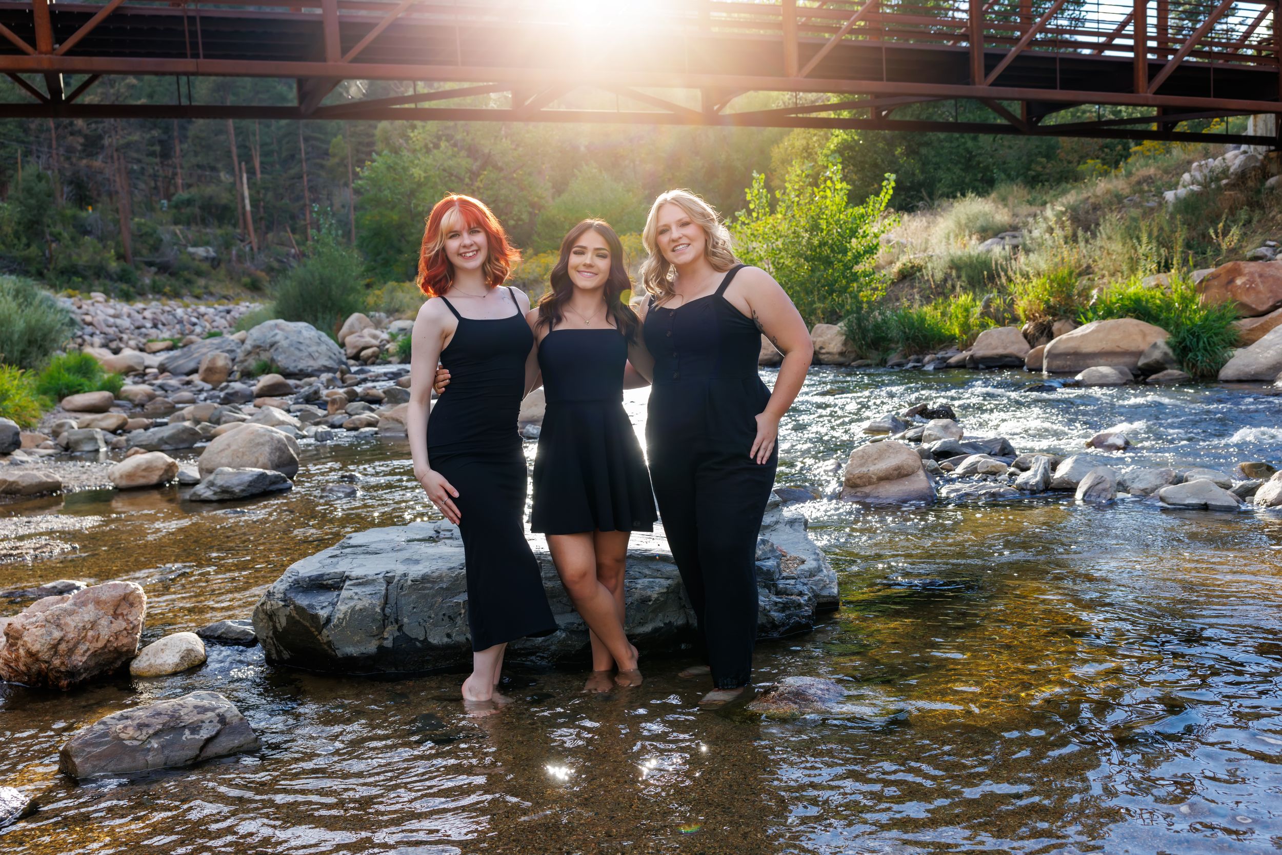 Three women in black dresses pose gracefully on rocks in a shallow creek beneath a rustic bridge at sunset.