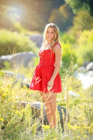 Young blonde woman in red sundress smiling outdoors in sunlit meadow with yellow wildflowers and rocks.