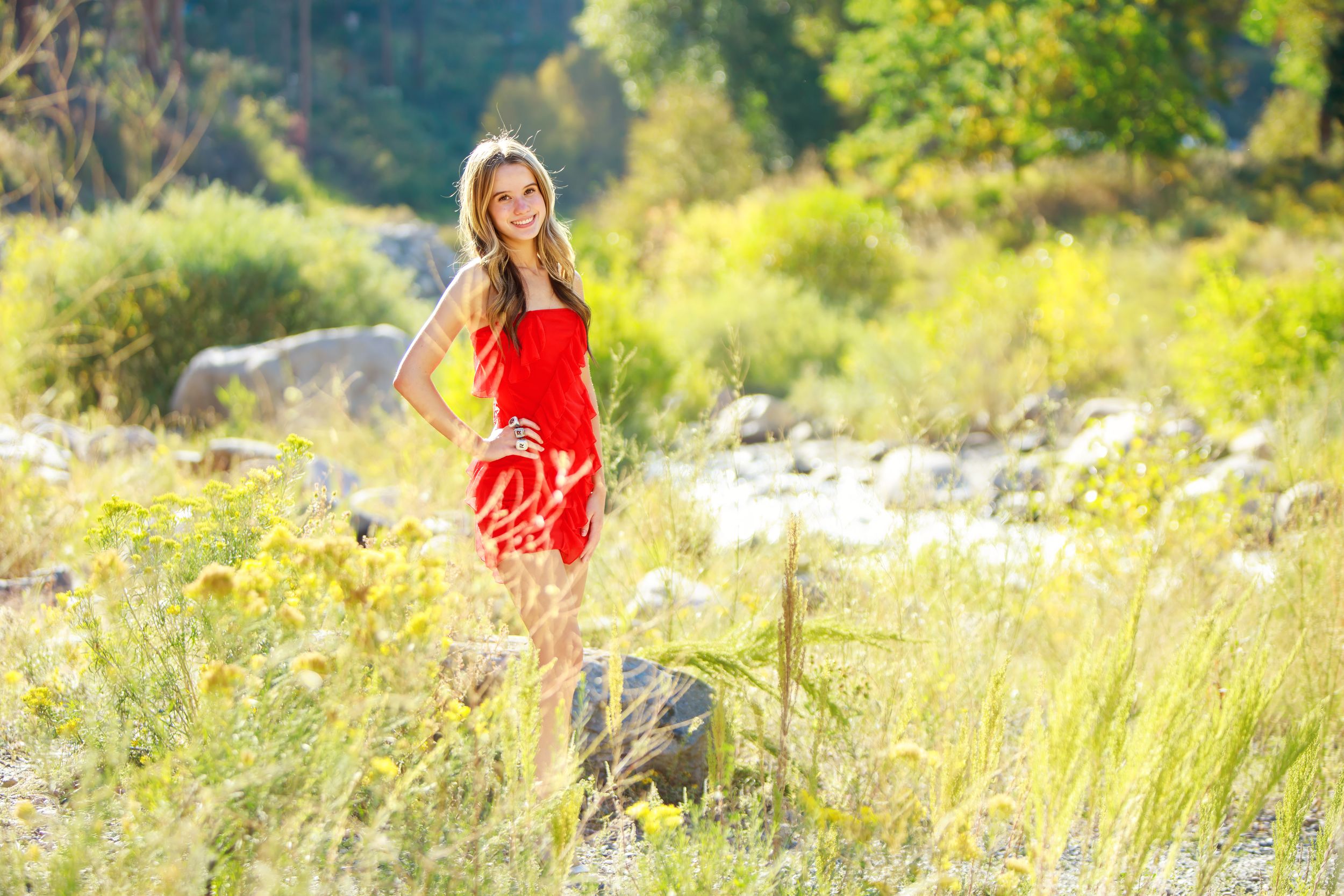 Woman in red sweater walking through a sunlit meadow with tall grass and wildflowers during golden hour.