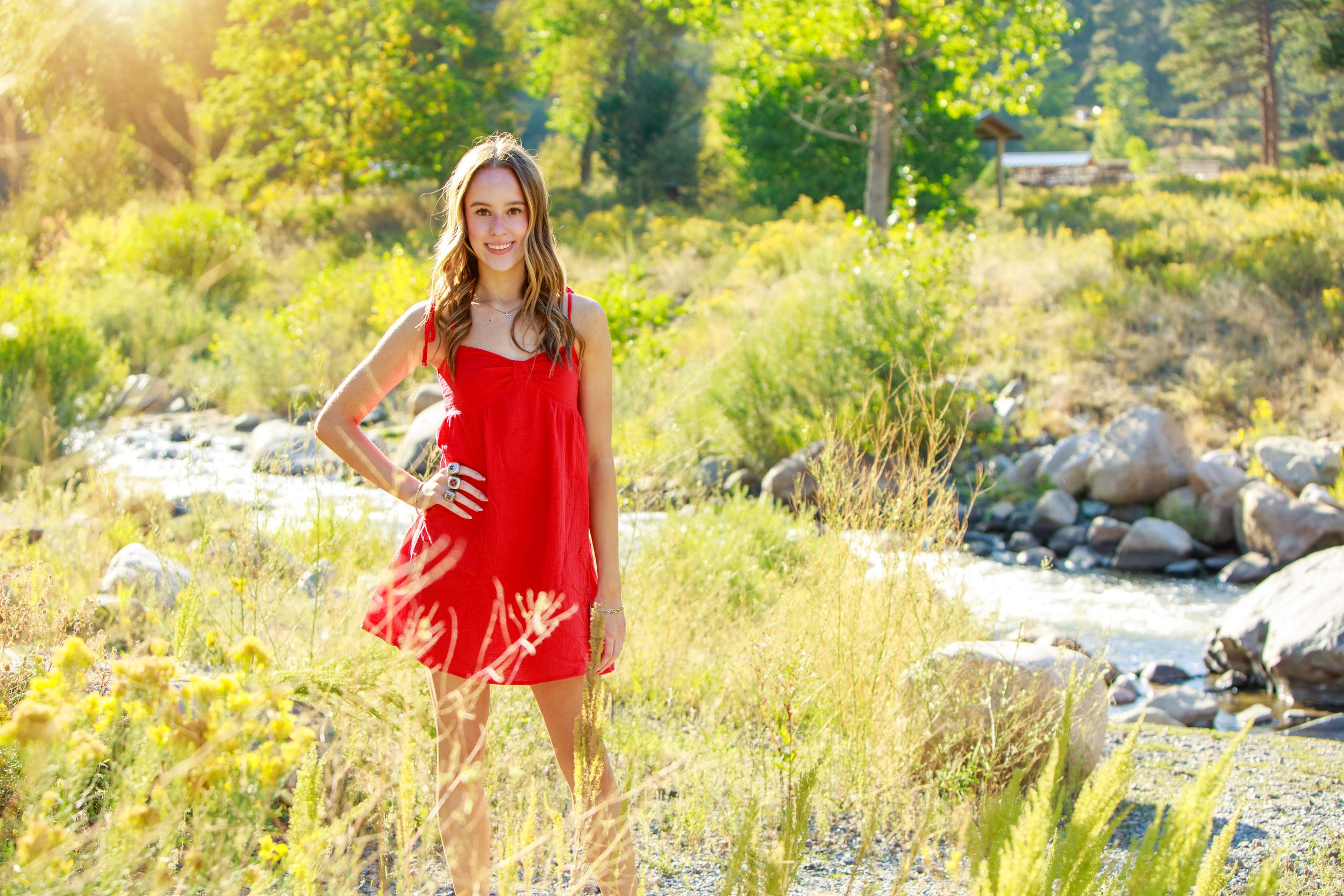 Young woman in red dress poses by a sunlit mountain stream surrounded by lush green grass and rocks.