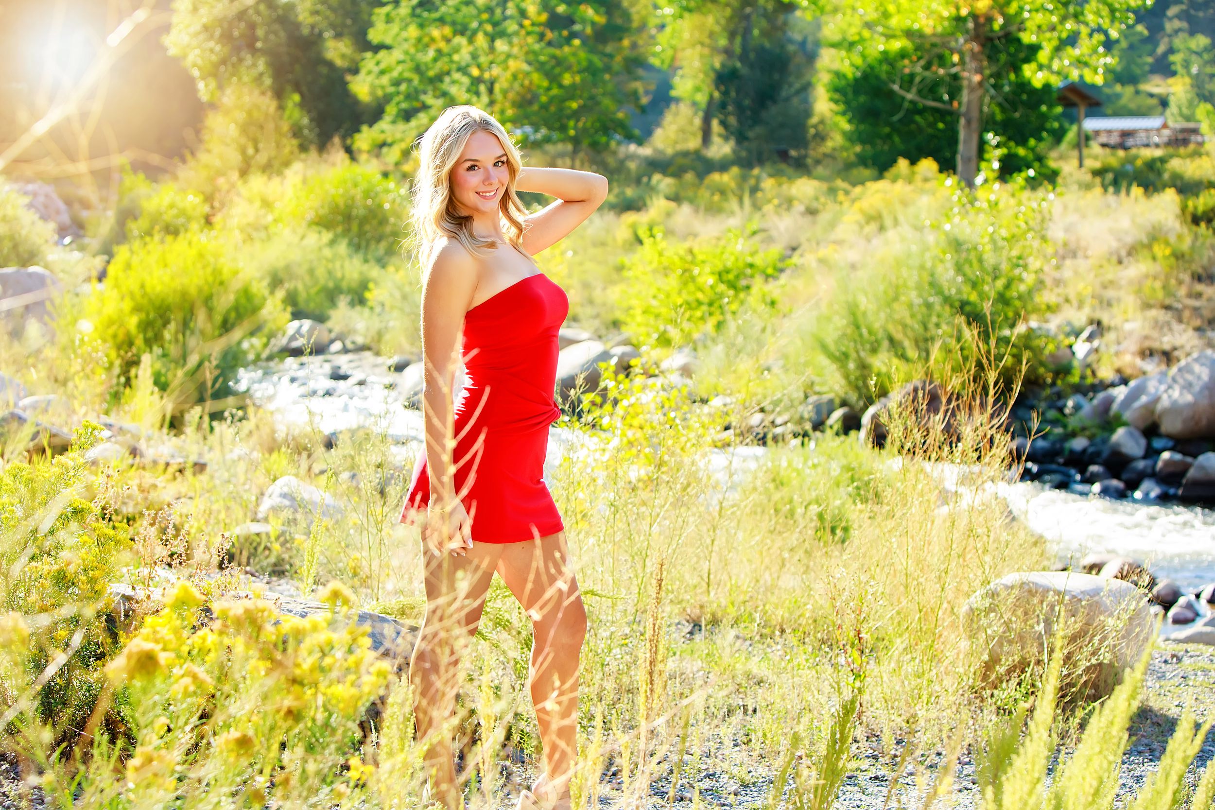 Blonde woman in red dress posing outdoors in sunlit natural setting with green foliage and rocky stream.