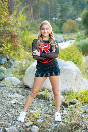 Smiling cheerleader in black and red uniform with letter R poses arms crossed on rocky riverbank surrounded by autumn trees.