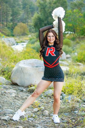 Cheerleader in black and red uniform with pom-poms poses outdoors on rocky terrain with lush greenery behind her.