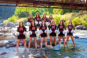 Cheerleading squad in black and red uniforms with white pom-poms posing by a rocky creek outdoors.