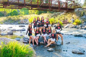 Group of girls in matching black and red uniforms posing on rocks by a mountain stream under a bridge.