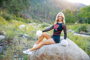Blonde cheerleader in black and red uniform poses on a boulder in a scenic mountain landscape.