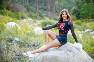 Cheerleader in black and red uniform with pom-poms poses on a rock surrounded by lush greenery outdoors.