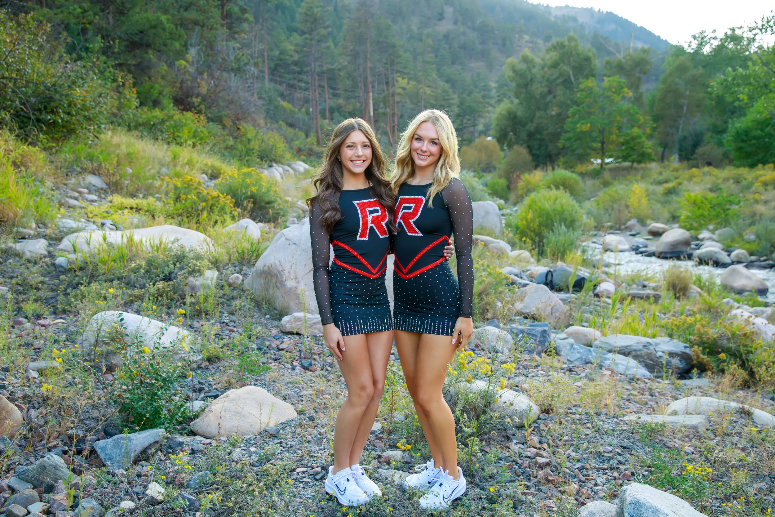 High school cheerleaders in red and black uniforms pose together on rocky terrain during an outdoor photo session.