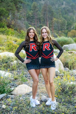 Two cheerleaders in matching black and red uniforms with letter R pose outdoors among rocks and wildflowers.