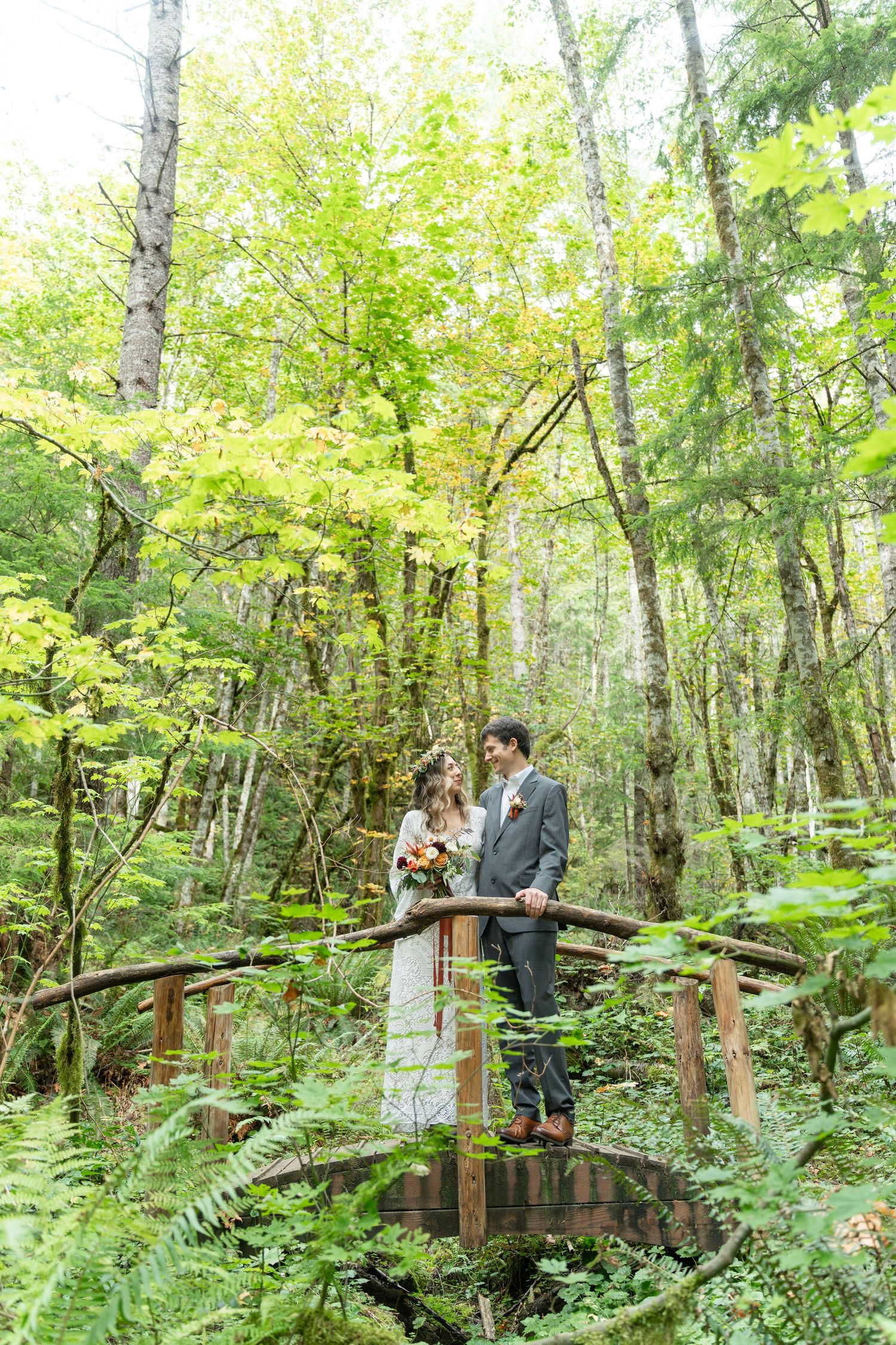 A couple stands together on a wooden footbridge surrounded by lush green forest and ferns in a romantic woodland setting.