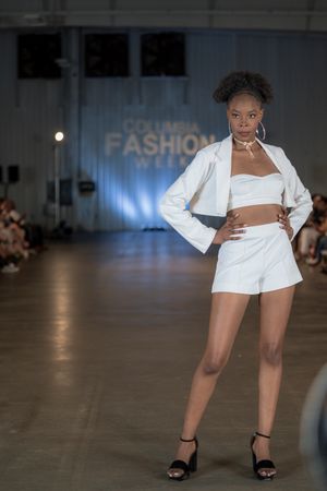 Fashion model showcases white blazer dress with black sandals on catwalk.