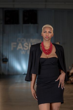Model wearing black strapless dress with blazer and striking red beaded necklace on fashion runway.