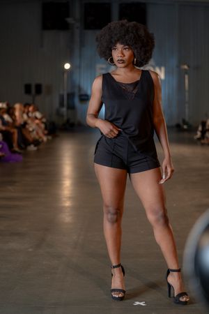 Fashion model showcases black romper with confidence on dimly lit catwalk.