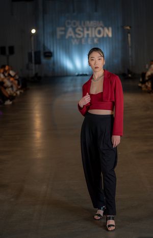 Model wearing red cropped jacket with black pants walks runway at fashion show.