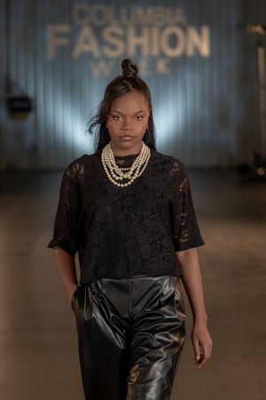 Model wearing black lace dress with pearl necklace walks fashion runway.
