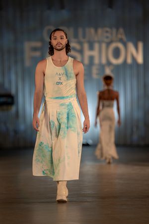 Model presents white and turquoise tie-dye outfit on fashion runway with blue backdrop.