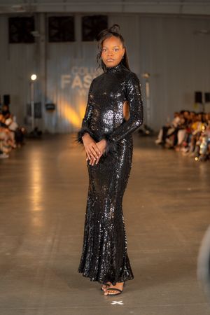 Model walks runway in sparkling black sequined gown with long sleeves at fashion show.