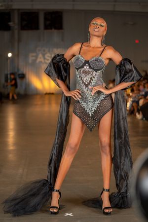 Model showcases dramatic black and silver corset gown with flowing cape on fashion runway.
