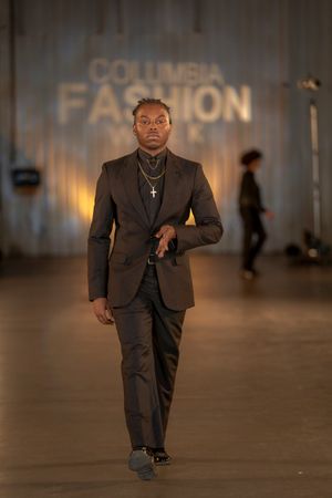 A suited model walks the runway in a dark tailored ensemble with cross necklace at Columbia Fashion Week.