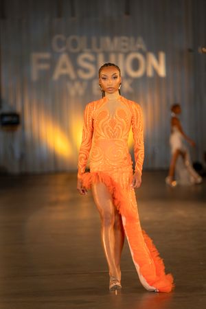 Model walks runway in flowing orange gown with dramatic train during fashion show presentation.