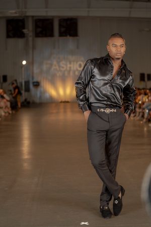 Model walks runway in black satin jumpsuit during fashion show with dramatic lighting.