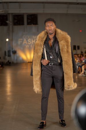 Model showcases black outfit with tan fur coat on fashion runway.
