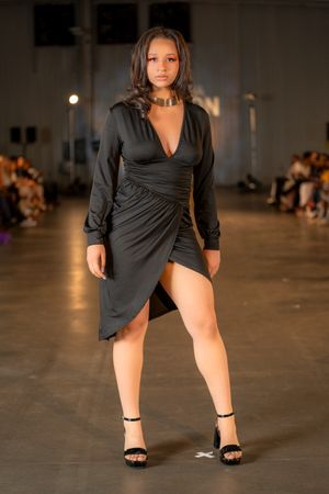Model wearing black wrap dress and strappy sandals poses on fashion runway.
