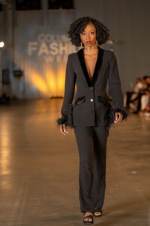 Model showcases elegant black pantsuit ensemble on fashion runway with warm lighting.