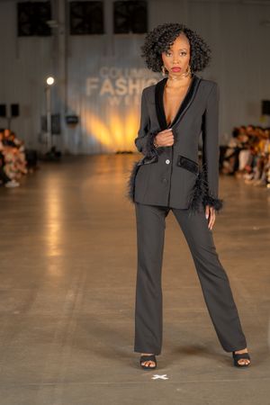 Professional model walks runway in charcoal grey pantsuit during fashion show.