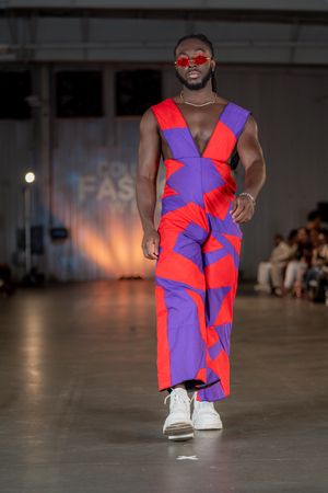 Model presents vibrant red and purple geometric jumpsuit with white sneakers on runway.