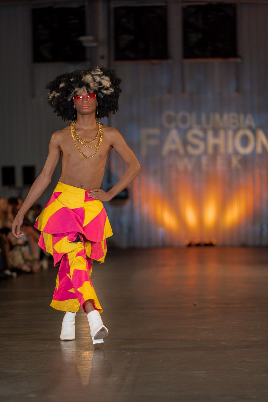 Model struts in vibrant pink and yellow ruffled outfit with white boots on fashion catwalk.