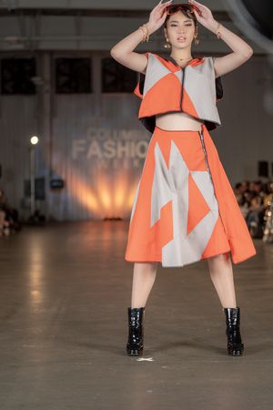 Model showcases geometric orange and white dress with black boots on fashion runway.