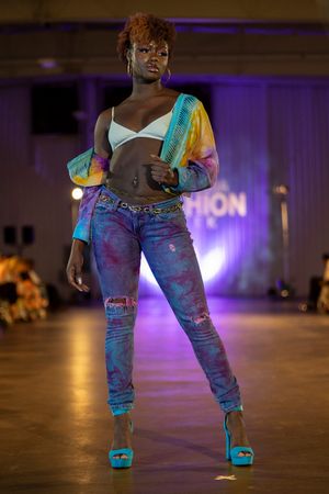 Model walks runway in distressed jeans, white crop top and colorful tie-dye jacket at fashion show with purple lighting.