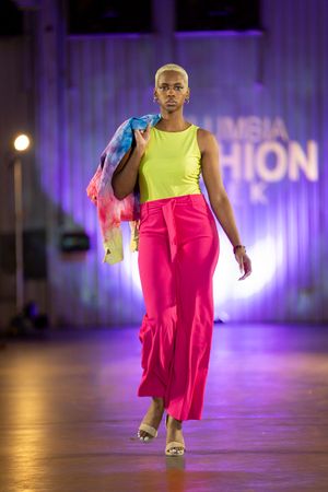 Fashion model wearing bright neon yellow top and hot pink skirt walks runway under purple stage lighting.